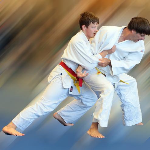 Zwei Judokas in Weiß kämpfen, einer greift den anderen an, während er nach vorne gedrängt wird. Hintergrund unscharf.