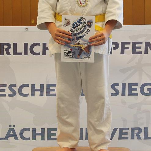 Junge in judogee mit Medaille und Urkunde auf einem Siegerpodest in einer Sporthalle.