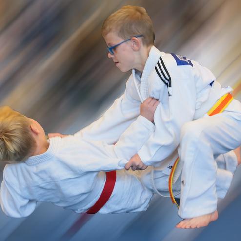 Zwei Kinder im Judoanzug führen einen Wurf aus, während sie auf einer unscharfen Fläche stehen.