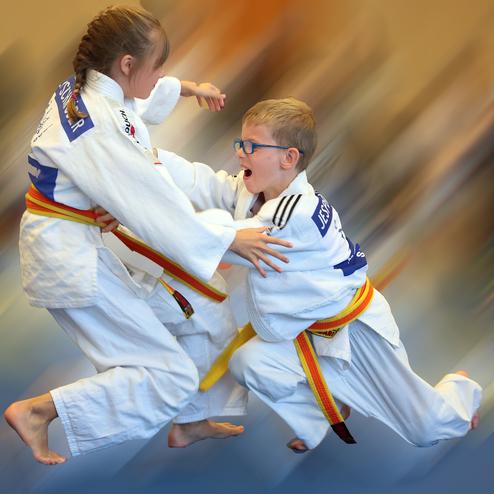 Zwei Kinder im Judoanzug kämpfen begeistert miteinander auf einer blauen Matte. Bewegung und Dynamik sind deutlich sichtbar.