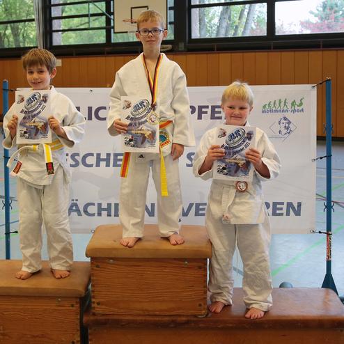 Drei Kinder in Karateanzügen stehen auf Podesten und halten Urkunden. Dahinter eine Banner mit Aufschrift.