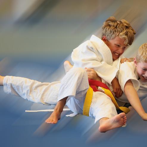 Zwei Kinder in weißen Judoanzügen spielen auf einer blauen Matte, eines hält das andere fest umarmt.