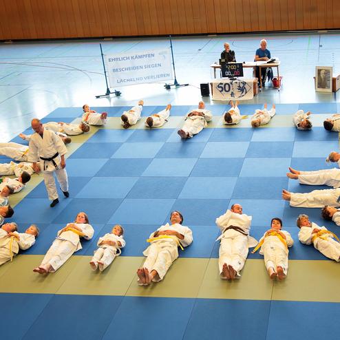 Gruppenübung von Judokas in weißen und gelben Anzügen auf blauer Matte; Lehrer leitet die Einheit.
