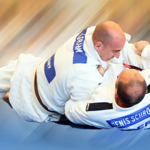 Zwei Judokas in weißen Anzügen kämpfen auf einer Matte, dynamische Bewegung und Konzentration sind deutlich erkennbar.