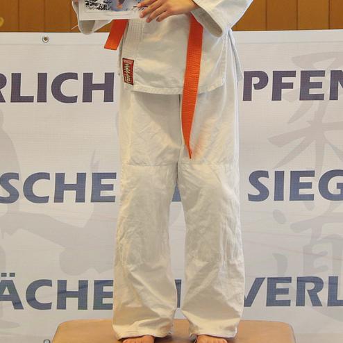 Mädchen in einem weißen Judogi mit Medaille und Urkunde auf einem Siegertreppchen in einer Sporthalle.