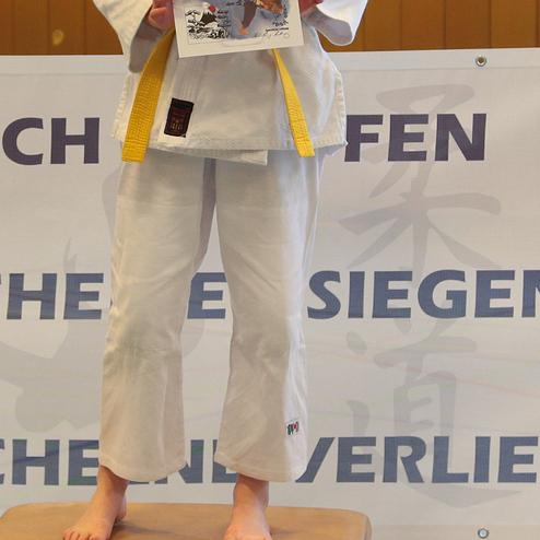 Mädchen in Karate-Uniform steht auf einem Podest und hält eine Urkunde bei einer Wettkampfveranstaltung.
