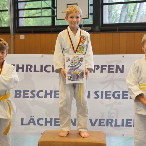 Drei Kinder in Judogi stehen auf dem Treppchen, halten Urkunden und tragen Medaillen in einer Sporthalle.