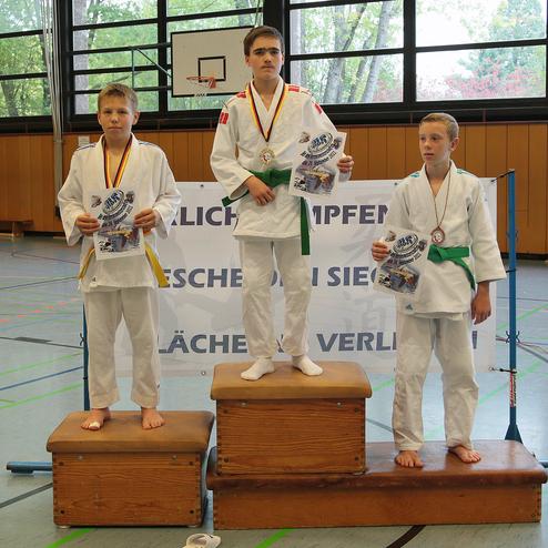 Drei Kinder in Karateanzügen stehen auf Podesten und halten Urkunden des Wettkampfs in einer Sporthalle.