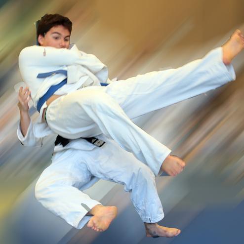 Jugendlicher in einem weißen Judoanzug führt einen Sprungkick inmitten einer verschwommenen Umgebung aus.