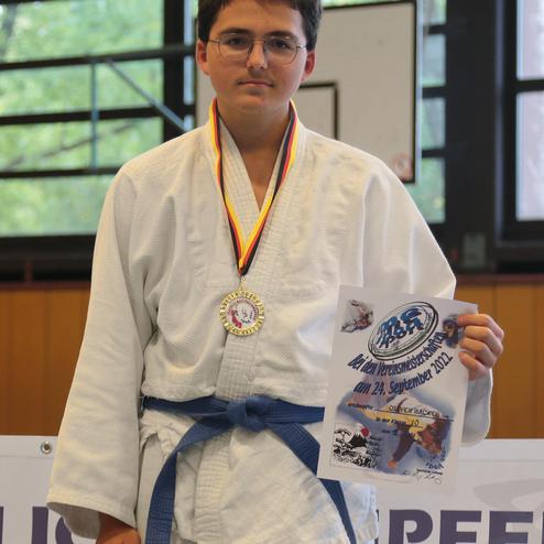 Junger Jude in Uniform mit blauer Gürtelschleife, hält eine Urkunde und trägt eine Medaille. Hintergrund zeigt Sporthalle.