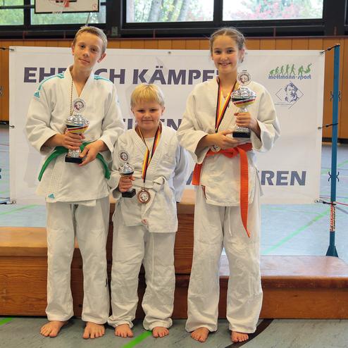 Drei Kinder in Judogi halten Pokale vor einer Wettkampfwand in einer Sporthalle.