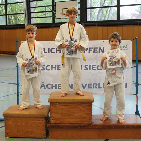 Drei Kinder in weißen Judoanzügen stehen auf Treppchen, halten Urkunden und Medaillen in einer Sporthalle.