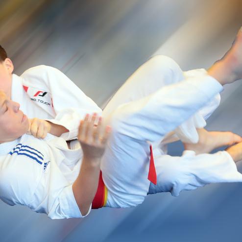 Zwei Jungs in weißen Judogis kämpfen; einer führt einen Wurf aus, während der andere festgehalten wird.