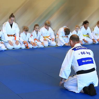 Gruppe von Judokas in weißen Anzügen sitzt auf blauen Matten und führt eine neue Übung durch.