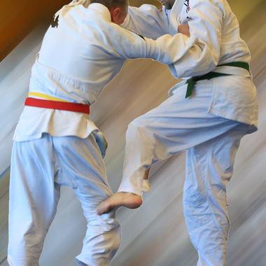 Zwei Judoka in weißen Gis sind im Wettkampf; einer hebt das Knie zur Ausführung eines Hebels.