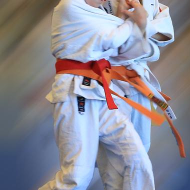 Zwei Mädchen in weißen Judogis mit roten Gurten führen einen Sprung im Judo aus. Hintergrund mit dynamischem Farbverlauf.