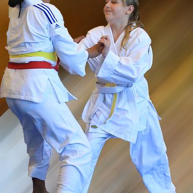 Zwei Kinder im Judoanzug üben einen Griff in einem Trainingsraum mit farbigen Gürteln.