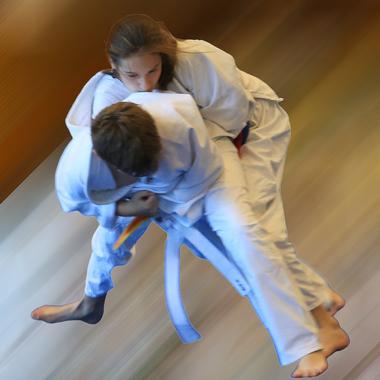 Zwei Kinder in wei&szlig;en Judoanz&uuml;gen f&uuml;hren einen Wurf im Judo auf einer unscharfen, bewegten Fl&auml;che aus.