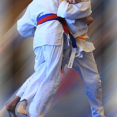 Zwei Kinder im Judo-Anzug umarmen sich während eines Kampfs, einer trägt einen roten, der andere einen gelben Gürtel.