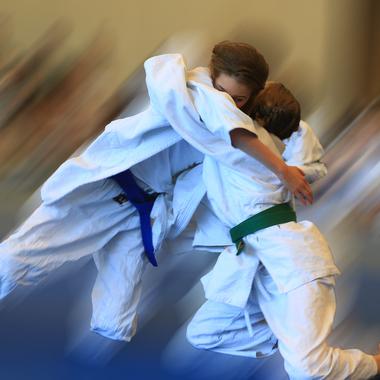 Zwei Judoka in Wei&szlig; und Gr&uuml;n k&auml;mpfen dynamisch auf einer blauen Judomatte, unscharf im Hintergrund Zuschauer.