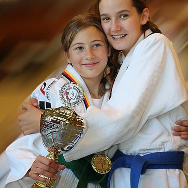 Zwei Mädchen in Judogi umarmen sich und halten einen Pokal sowie eine Medaille in der Hand, lächelnd in die Kamera.