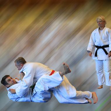 Zwei Judokas in Wettkampfsituation auf einer Matte, w&auml;hrend eine Trainerin im Hintergrund beobachtet.