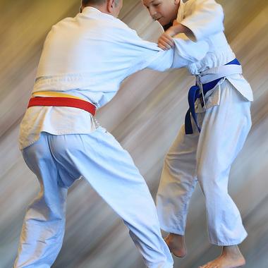 Zwei Judokas in weißen Anzügen üben einen Wurf, einer trägt einen blauen, der andere einen roten Gürtel.