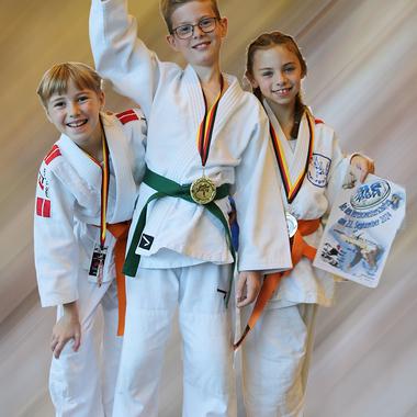 Drei Kinder in Judogi halten einen Pokal und zeigen Medaillen, sie stehen l&auml;chelnd vor einem verschwommenen Hintergrund.