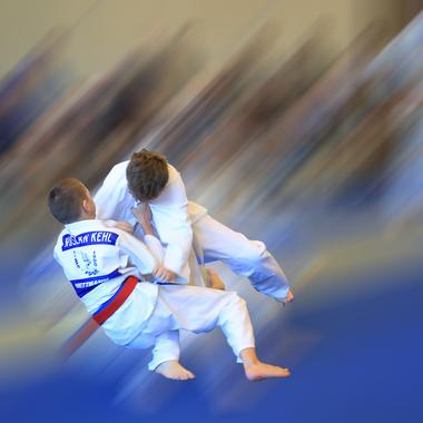 Zwei Kinder in wei&szlig;en Judoanz&uuml;gen f&uuml;hren einen Wurf auf einer blauen Judomatte aus, umgeben von verschwommenen Zuschauern.