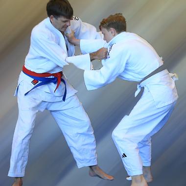 Zwei Judoka in wei&szlig;en Anz&uuml;gen k&auml;mpfen, einer mit rotem und der andere mit braunem G&uuml;rtel, auf verschwommener Hintergrundfl&auml;che.