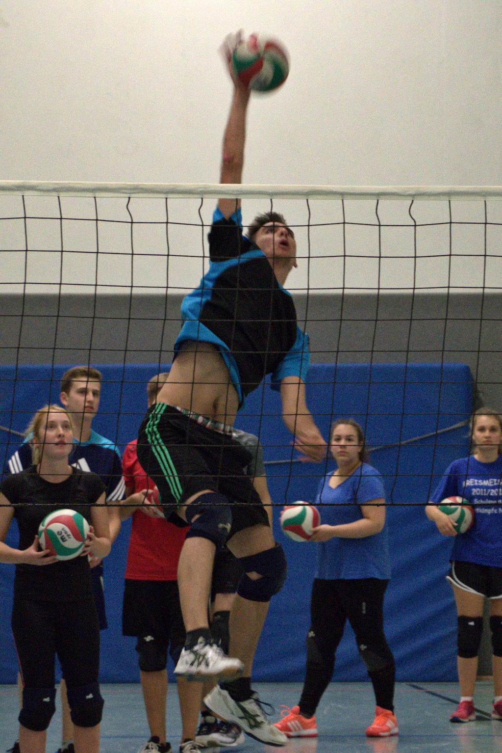 Ein Volleyballspieler springt für einen Aufschlag über das Netz, während andere Spieler im Hintergrund zuschauen.