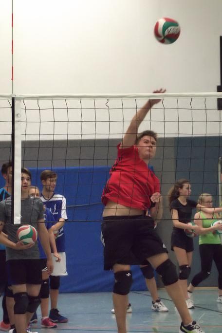 Junge in rotem Shirt springt, um einen Volleyball über das Netz zu schlagen, während andere Spieler im Hintergrund stehen.