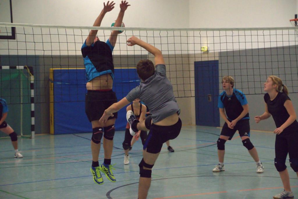 Zwei Volleyballspieler springen zum Blocken, während weitere Spieler auf dem Spielfeld in Aktion sind.