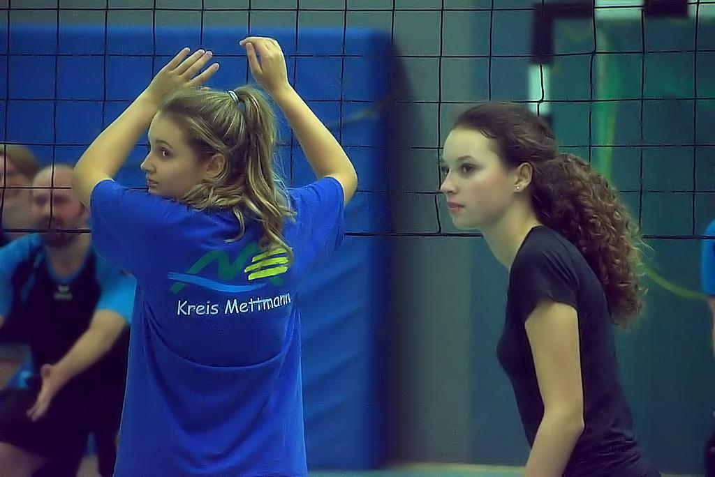 Zwei Mädchen in Sportkleidung stehen in einer Volleyballhalle, bereit für das Spiel hinter einem Netz.