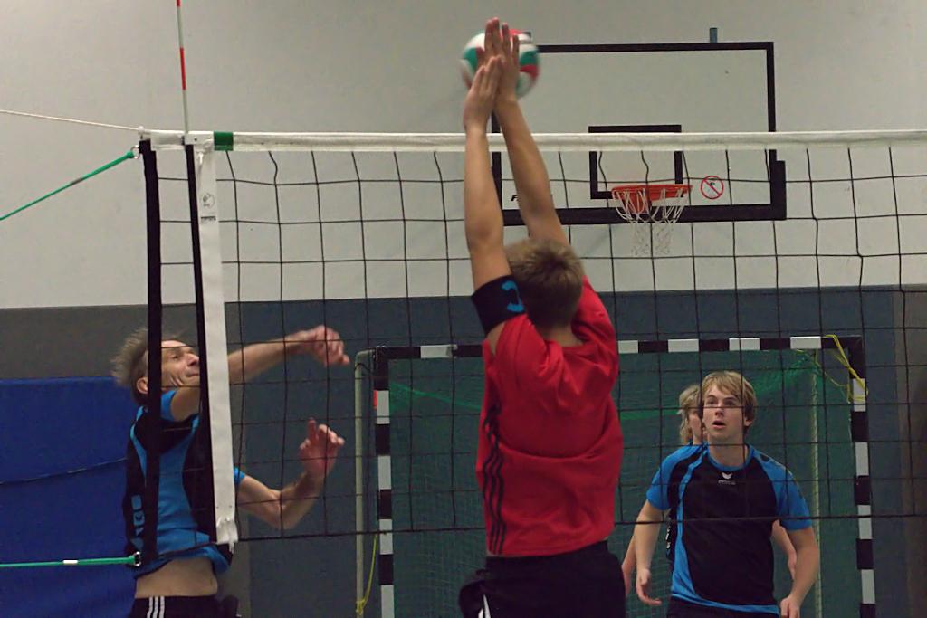 Zwei Spieler im Volleyball springen zur Ballabwehr am Netz, während ein weiterer Spieler beobachtet.