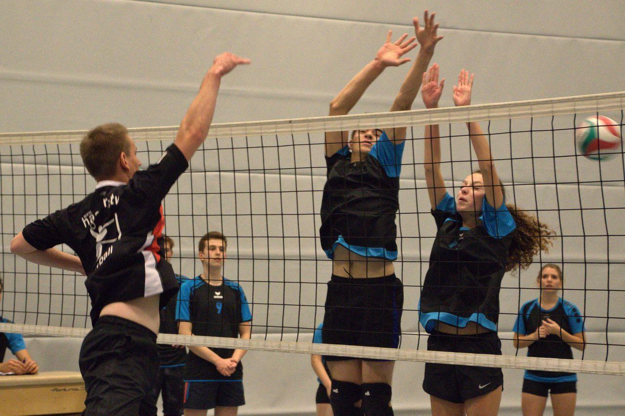 Zwei Spieler blocken einen Volleyball, während ein Angreifer den Ball über das Netz schlägt. Zuschauer im Hintergrund.