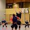 Volleyball-Spieler springen am Netz, während ein Ball in der Luft fliegt; weitere Spieler im Hintergrund beobachten.