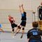 Zwei Volleyballspieler springen zum Ball, während ein Schiedsrichter auf einem Tisch steht und das Spiel beobachtet.