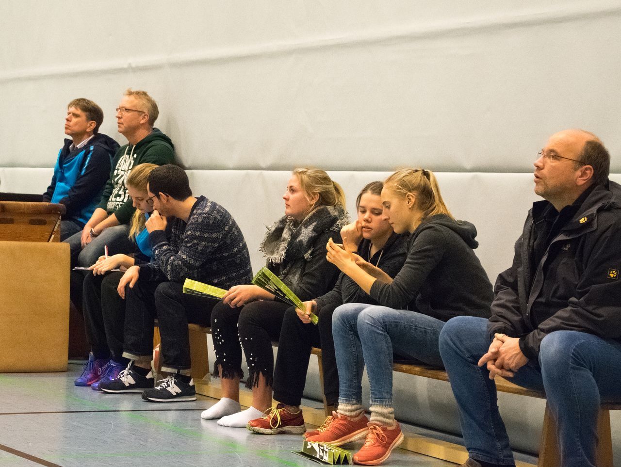 Eine Gruppe von sieben Personen sitzt auf einer Bank in einer Sporthalle und schaut in verschiedene Richtungen.