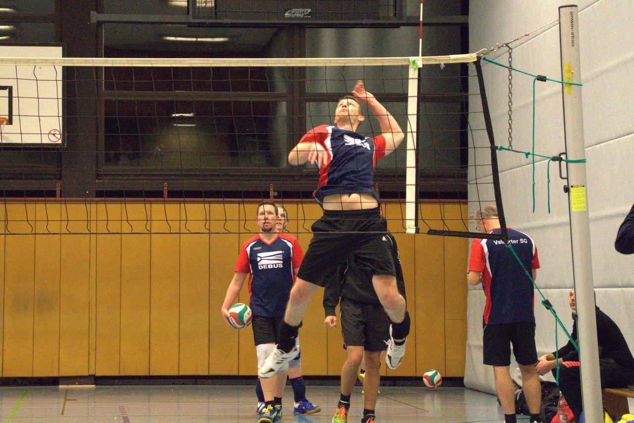 Spieler springt zum Blocken an einem Volleyballnetz in einer Sporthalle, andere Spieler stehen im Hintergrund.