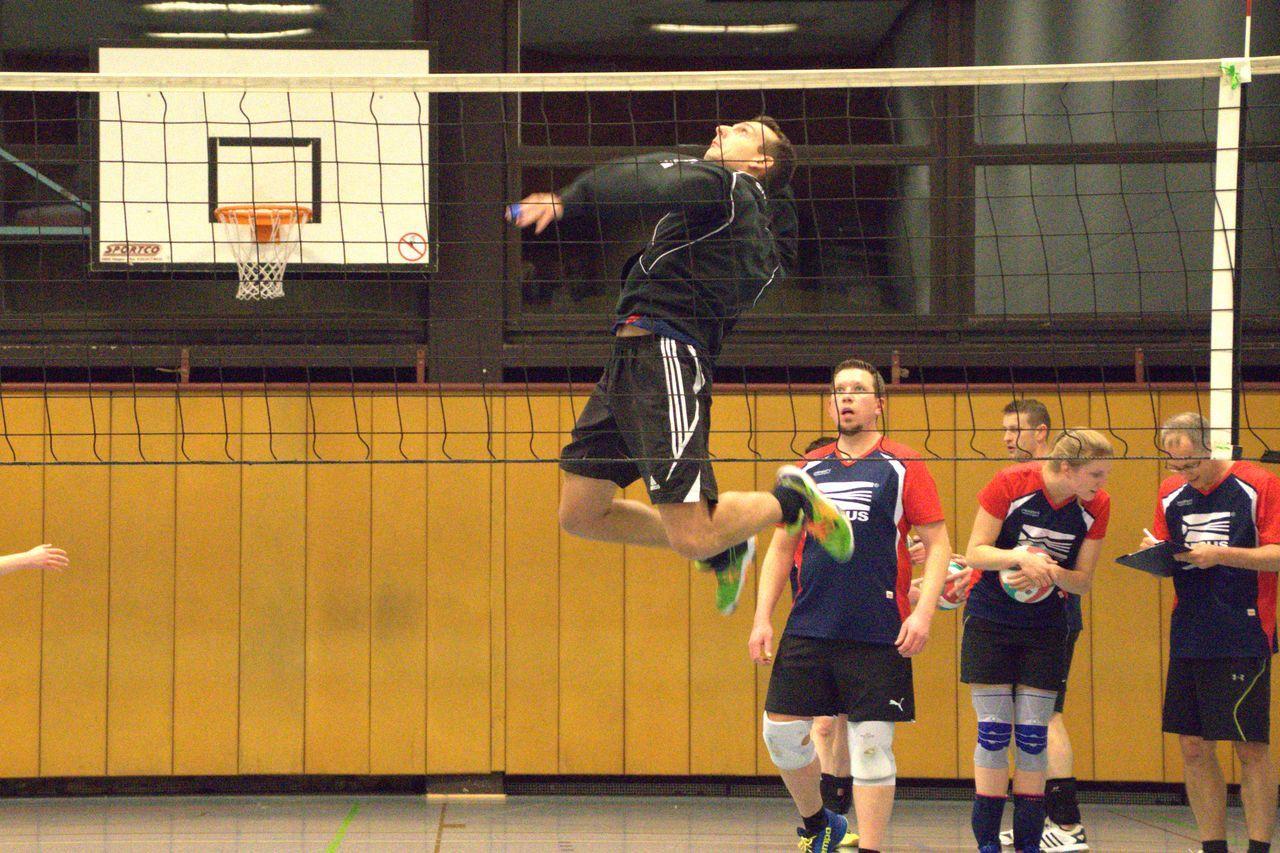 Ein Spieler springt zum Angriff über das Volleyballnetz, während Teamkollegen in sportlicher Kleidung zuschauen.