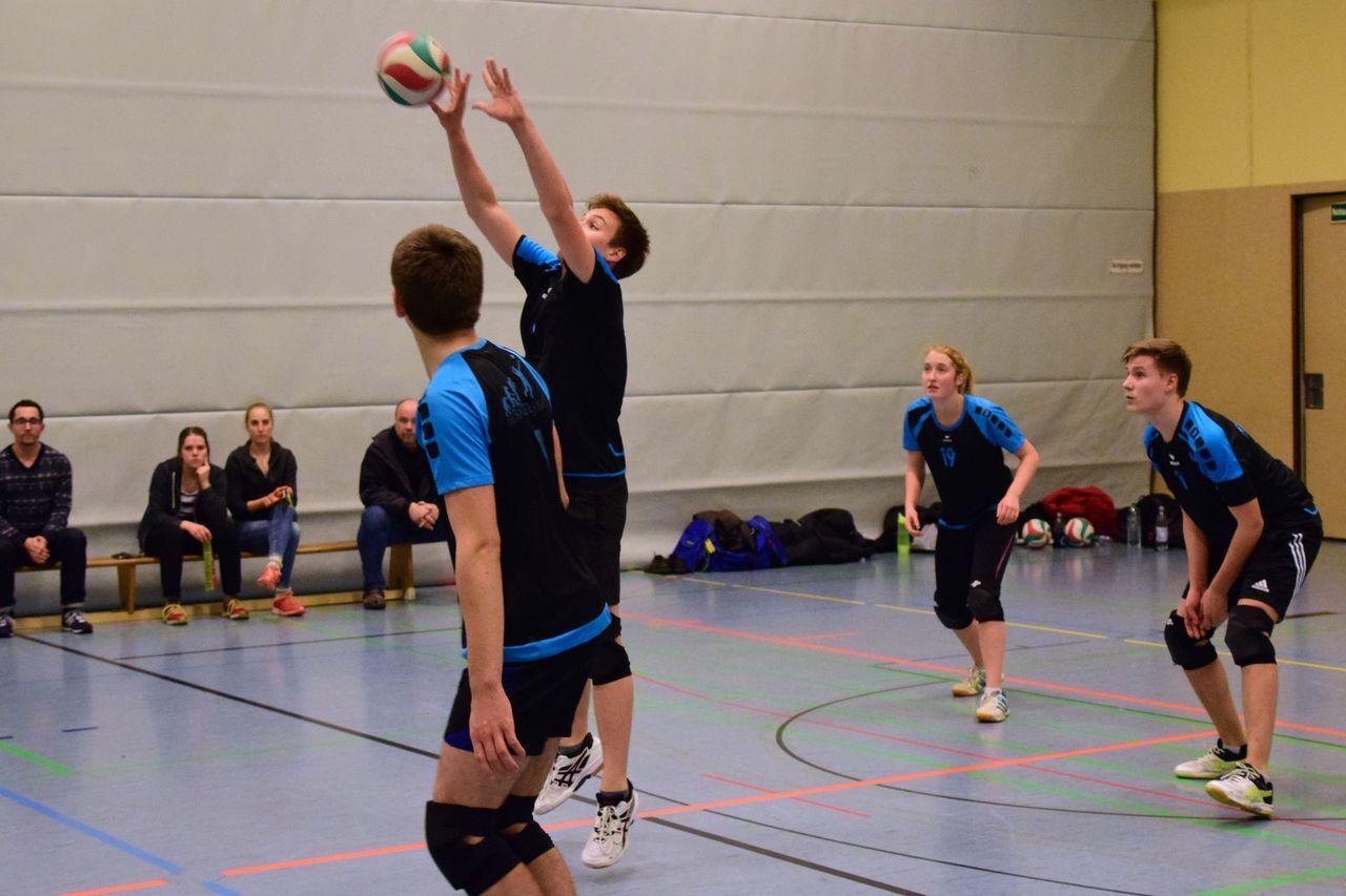 Ein Volleyballspieler hebt den Ball zum Zuspiel, während drei Mitspieler und Zuschauer im Hintergrund zu sehen sind.