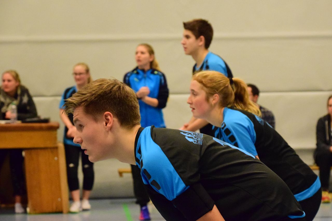 Drei Sportler in blauen Trikots in Spielposition, im Hintergrund weitere Teammitglieder in einer Sporthalle.