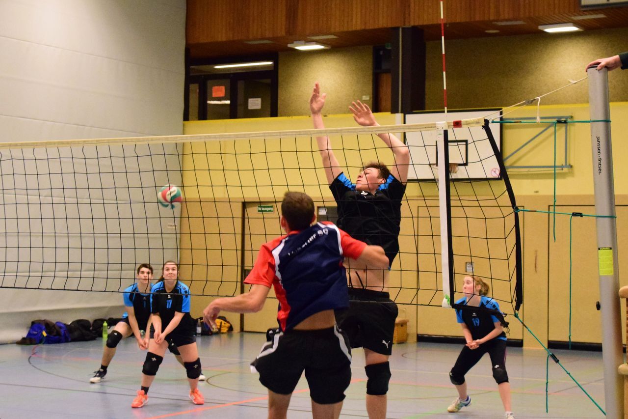 Volleyball-Spieler springen am Netz, während ein Ball in der Luft fliegt; weitere Spieler im Hintergrund beobachten.