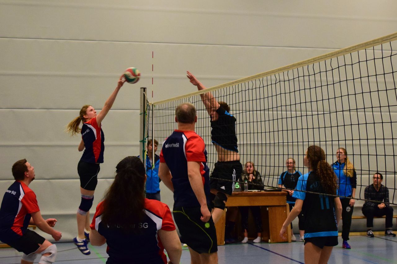Spielerinnen und Spieler beim Volleyball-Match, eine Spielerin springt zum Angriff über das Netz. Zuschauer im Hintergrund.
