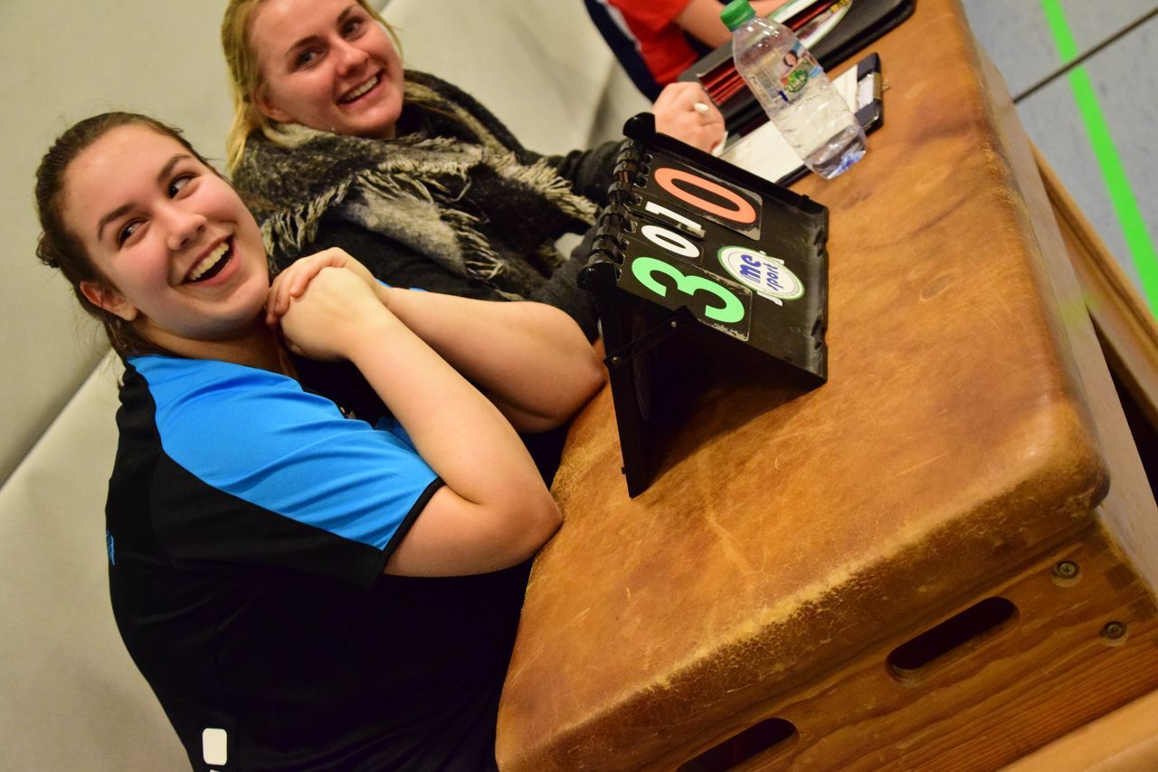 Zwei Frauen lächeln am Tisch mit einer Scoretafel, die den Punktestand 3:0 anzeigt. Wasserflasche und Unterlagen sind sichtbar.