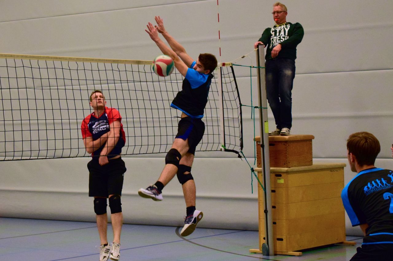 Zwei Volleyballspieler in Aktion, einer schlägt den Ball, während ein Schiedsrichter auf einer erhöhten Position beobachtet.