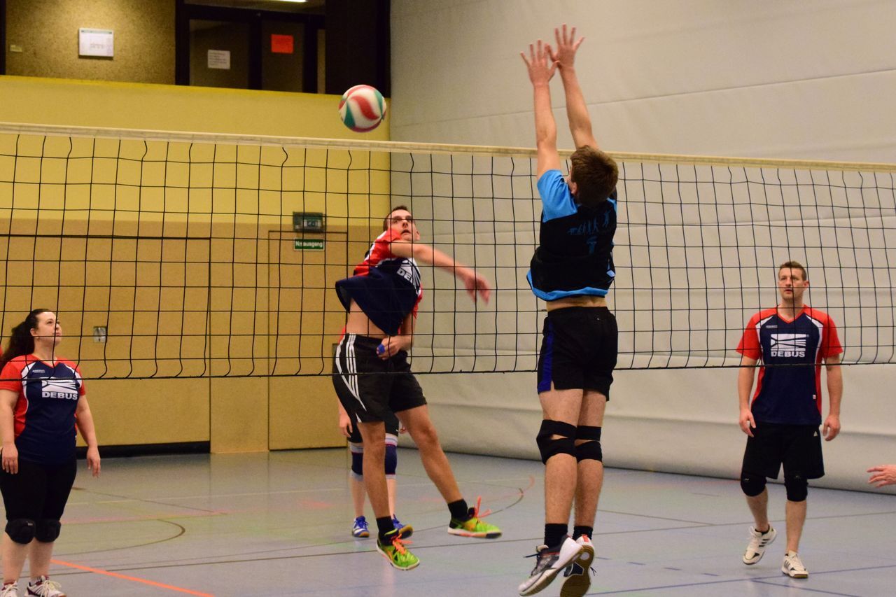 Zwei Volleyballspieler treffen sich am Netz, während eine Spielerin im Hintergrund zuschaut. Ein Ball schwebt darüber.