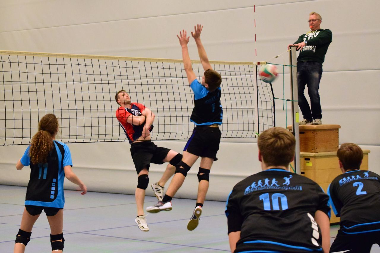 Zwei Volleyballspieler springen zum Ball, während ein Schiedsrichter auf einem Tisch steht und das Spiel beobachtet.