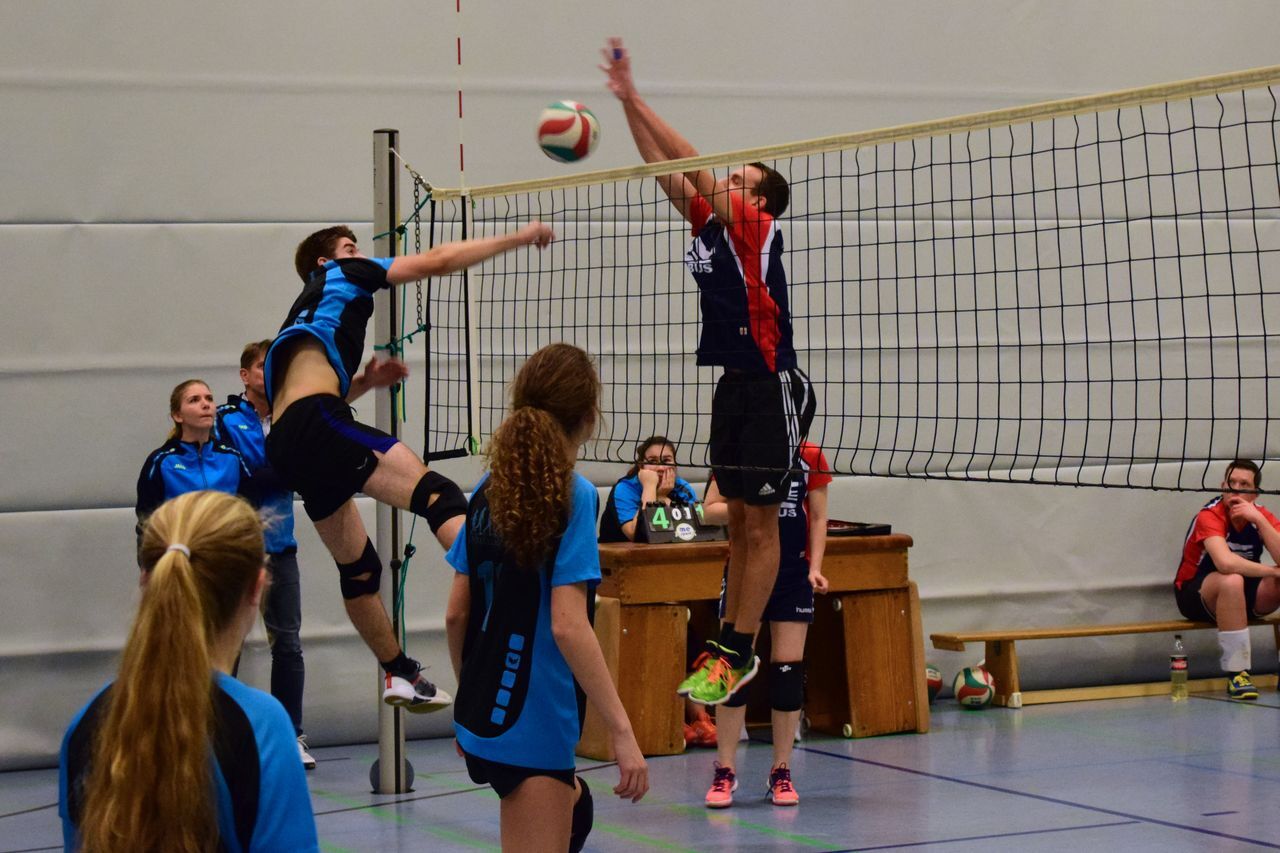 Zwei Volleyballspieler in Aktion beim Blocken am Netz, Zuschauer im Hintergrund, Sporthalle mit blauen Wänden.